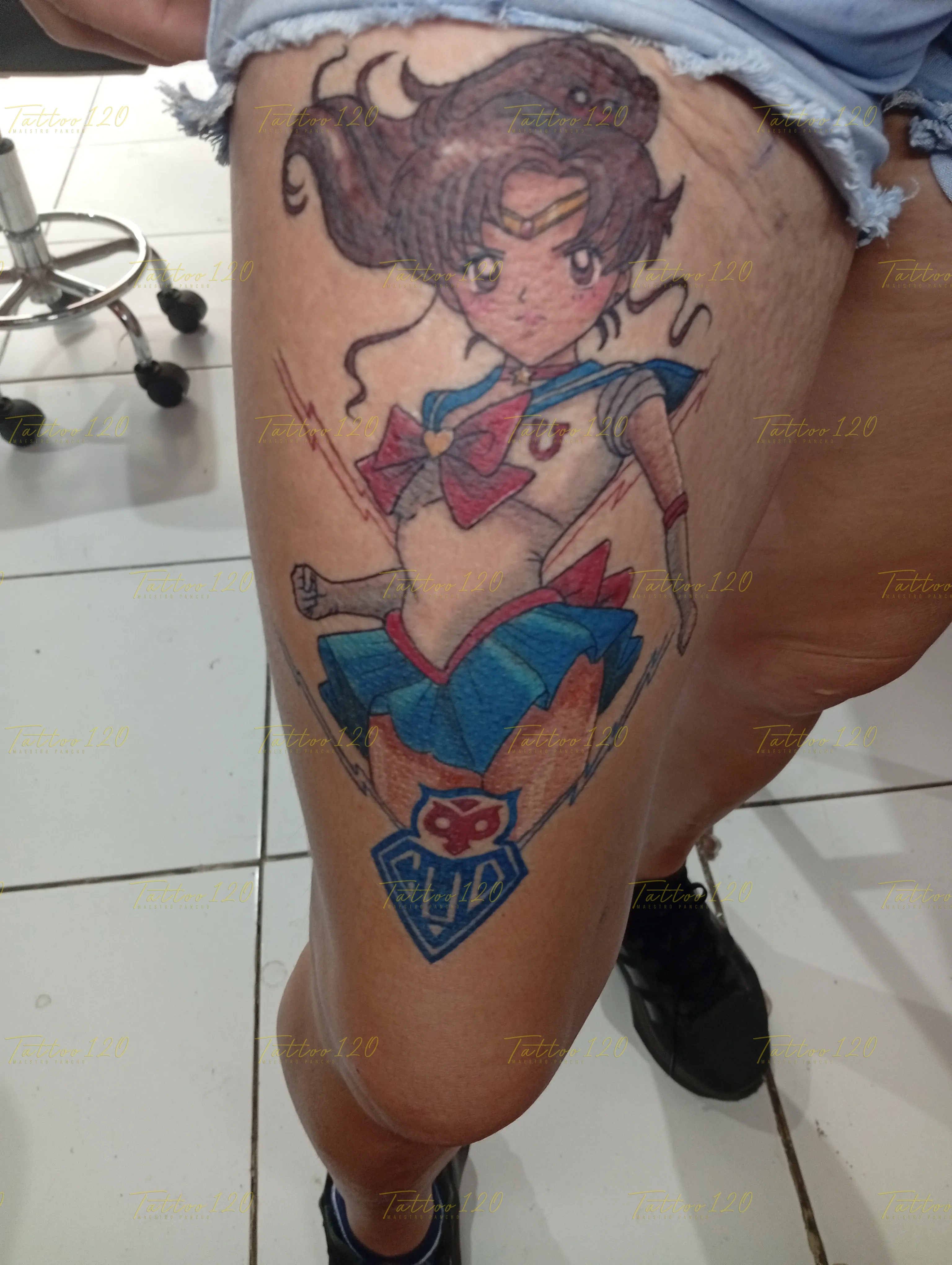 Tatuaje a color en el muslo de Sailor Jupiter de Sailor Moon en una pose dinámica, con un logotipo azul en la parte inferior sobre piel expuesta. estilo anime, manga e ilustraciones, realizado en el estudio Tattoo120 en Colina, Región Metropolitana.