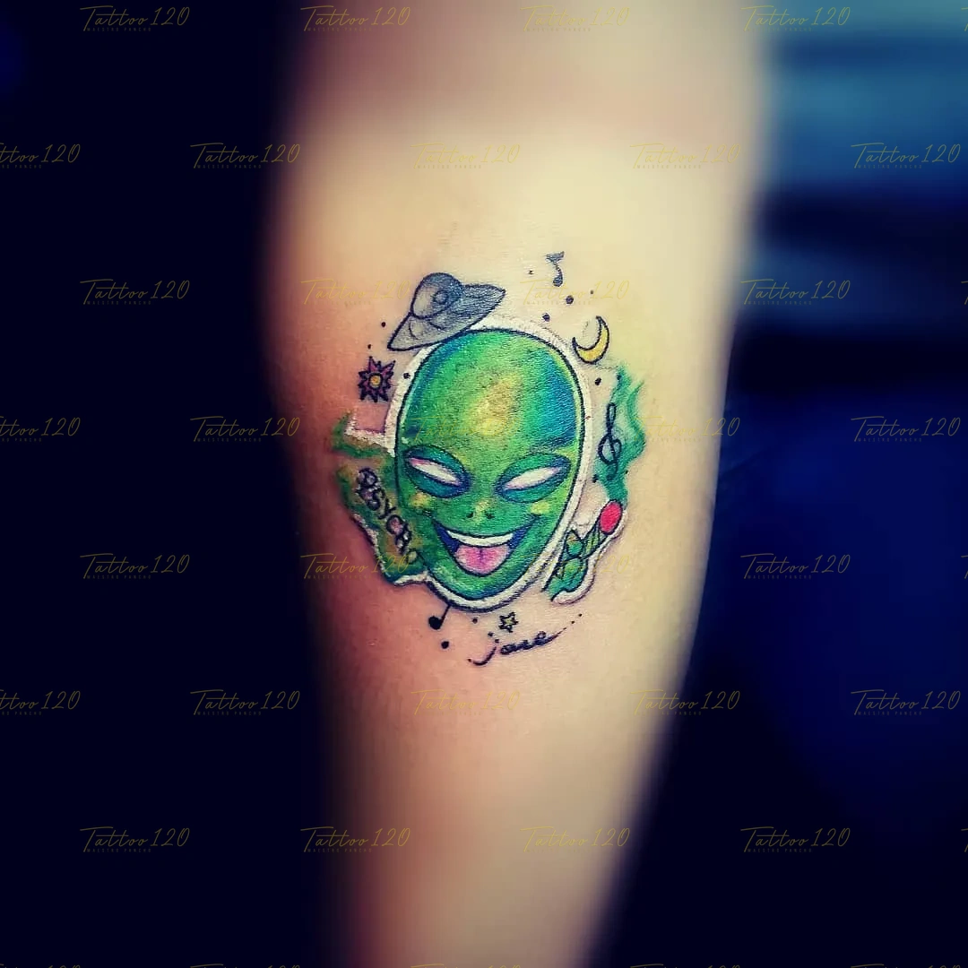 Tatuaje pequeño a color de la cabeza de un alienígena verde sacando la lengua, decorado con un ovni, notas musicales y una banda que dice 'PSYCHO'. estilo anime, manga e ilustraciones, realizado en el estudio Tattoo120 en Colina, Región Metropolitana.