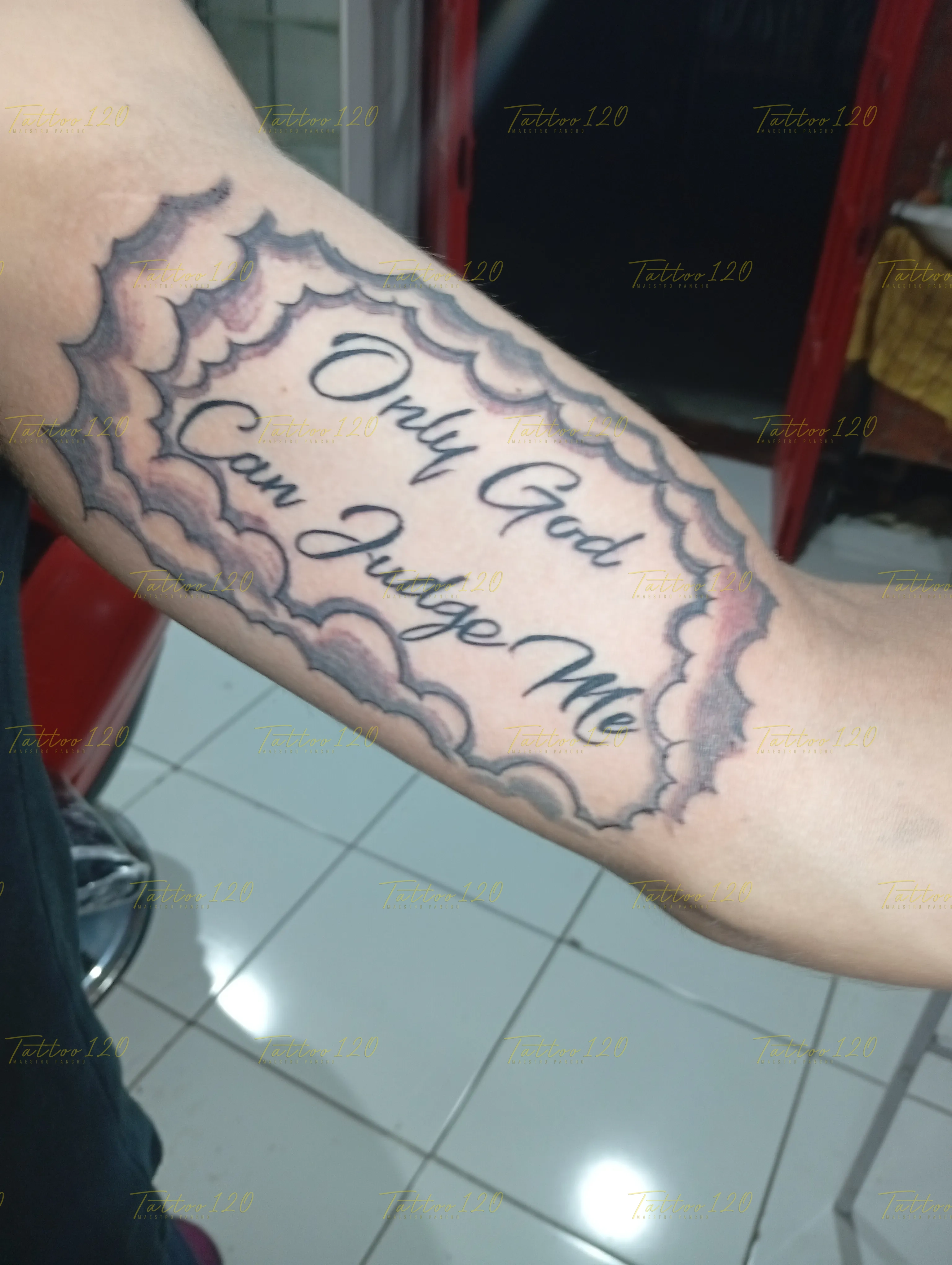 Tatuaje en el brazo con la icónica frase Only God Can Judge Me enmarcada por un fondo de nubes difuminadas, en estilo de letras, frases y chicano, realizado en el estudio Tattoo120 en Colina, Región Metropolitana.