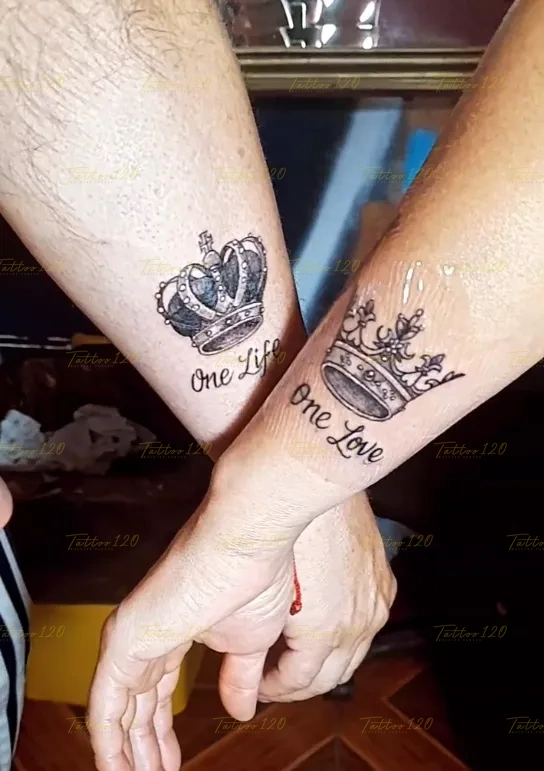 Tatuaje de parejas en las muñecas que muestra una corona de rey con la frase One Life y una corona de reina con One Love, en estilo de letras, frases y chicano, realizado en el estudio Tattoo120 en Colina, Región Metropolitana.
