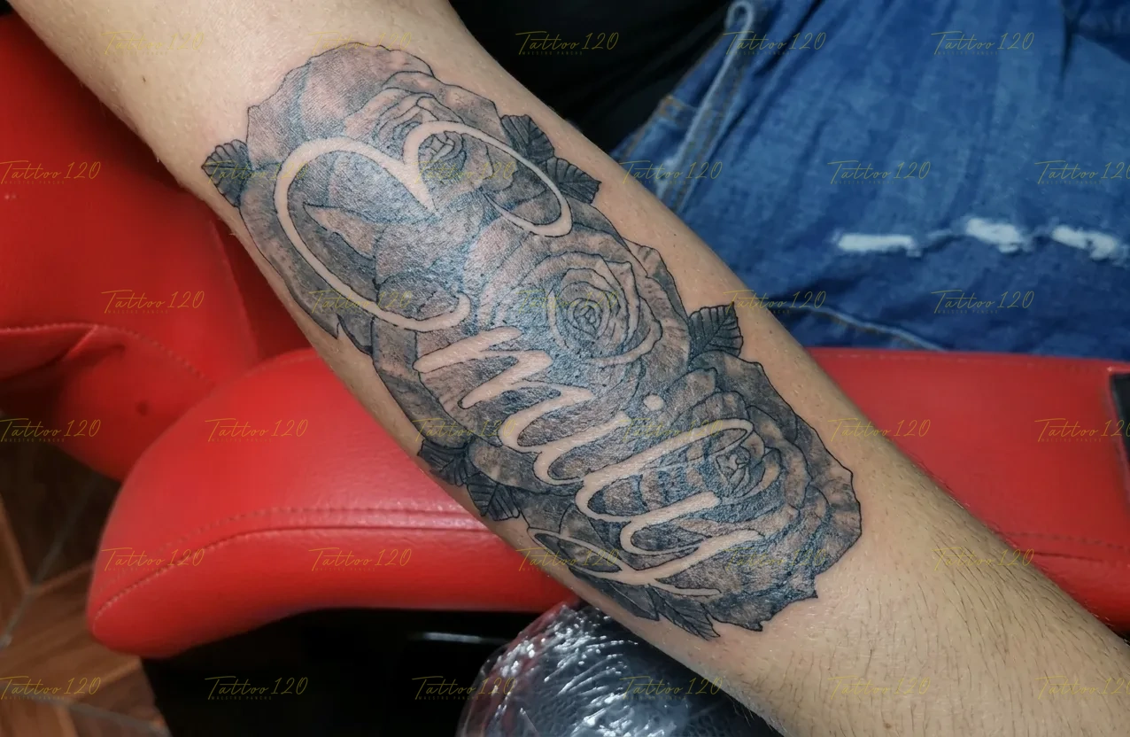 Tatuaje con el nombre Emily en letras blancas integradas de forma negativa sobre un fondo denso de rosas sombreadas, en estilo de letras, frases y chicano, realizado en el estudio Tattoo120 en Colina, Región Metropolitana.