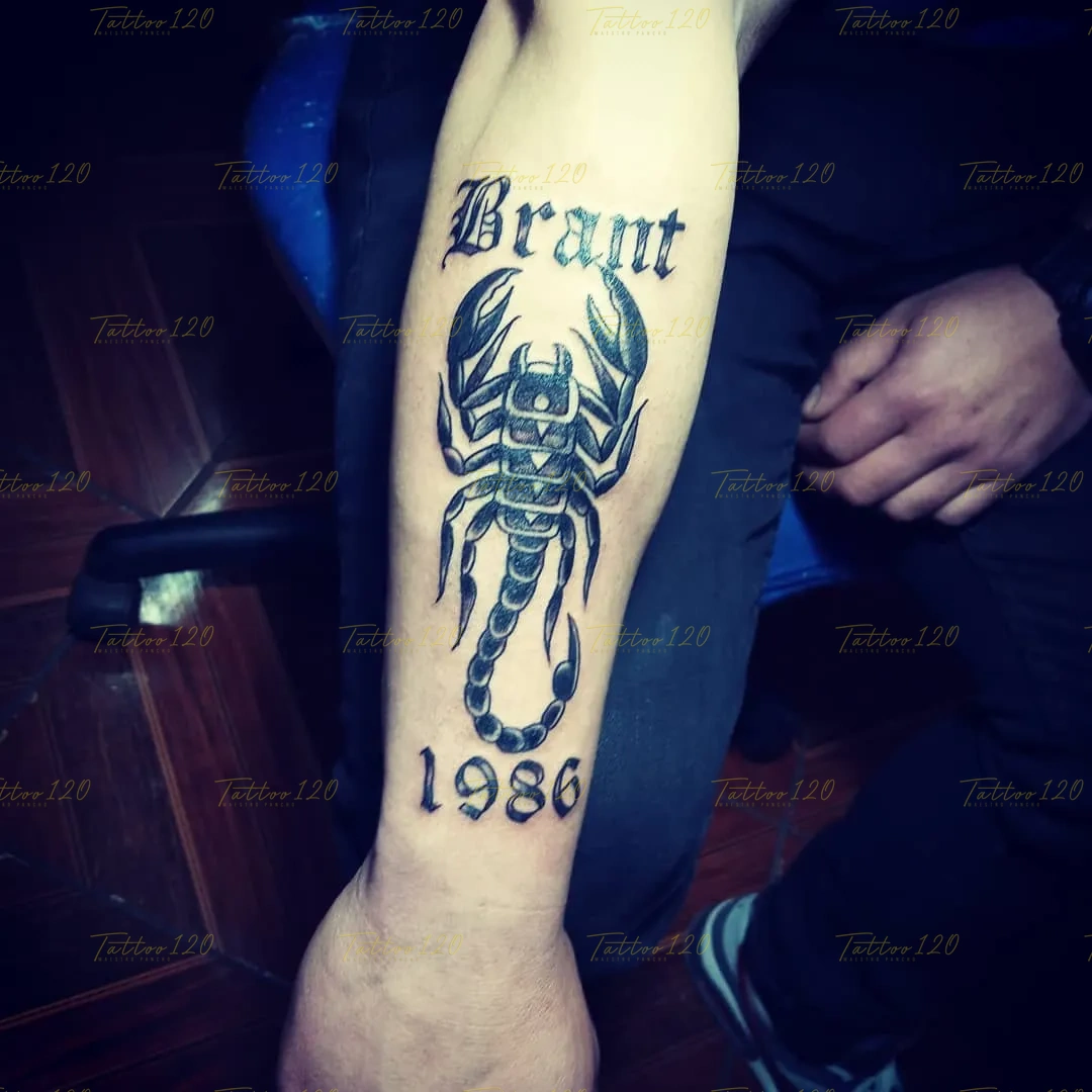 Tatuaje en el antebrazo que incluye el nombre Brant en letras góticas, un gran escorpión negro y el año 1986 abajo, en estilo de letras, frases y chicano, realizado en el estudio Tattoo120 en Colina, Región Metropolitana.