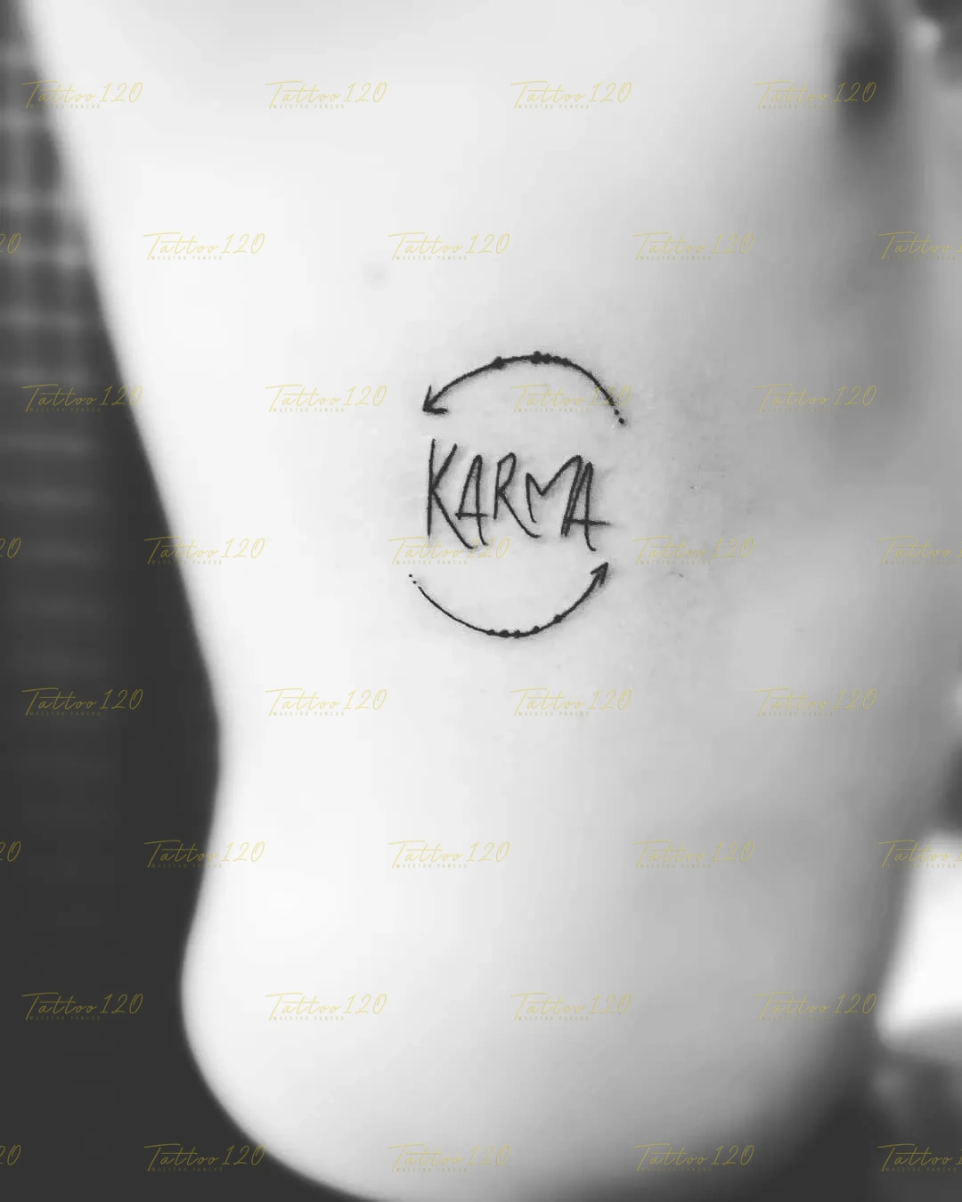 Tatuaje minimalista de la palabra Karma escrita con tipografía gestual y encerrada entre dos flechas circulares en la espalda, en estilo de línea fina y minimalistas, realizado en el estudio Tattoo120 en Colina, Región Metropolitana.