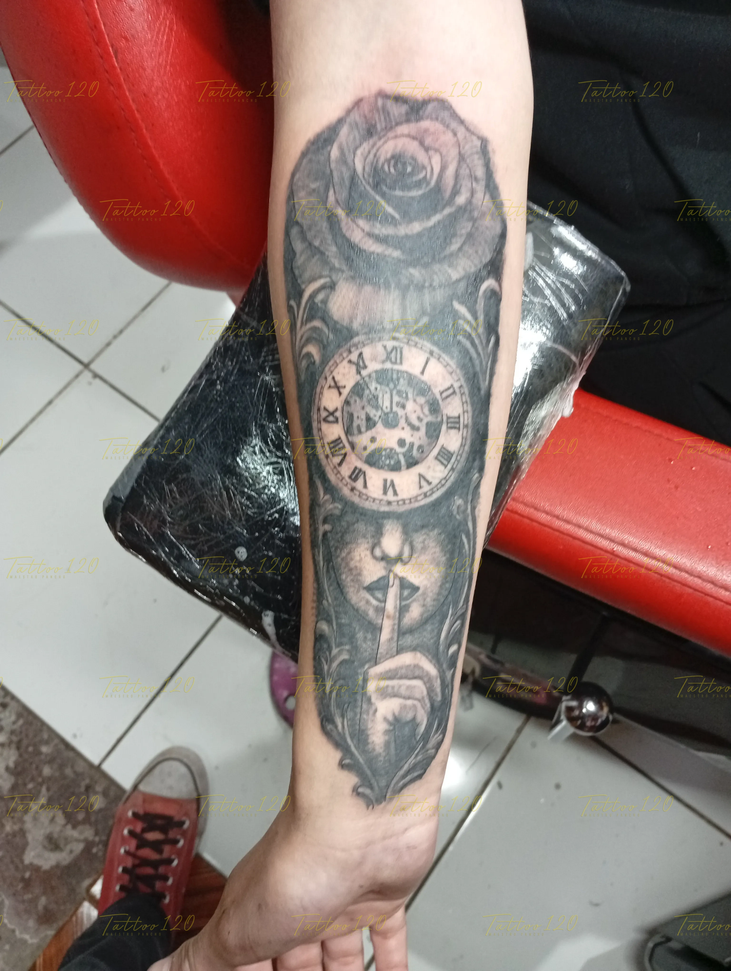 Tatuaje en el antebrazo que muestra la mitad del rostro de una mujer haciendo el gesto de silencio bajo un reloj mecánico y una rosa, en estilo de realistas, retratos y sombras, realizado en el estudio Tattoo120 en Colina, Región Metropolitana.