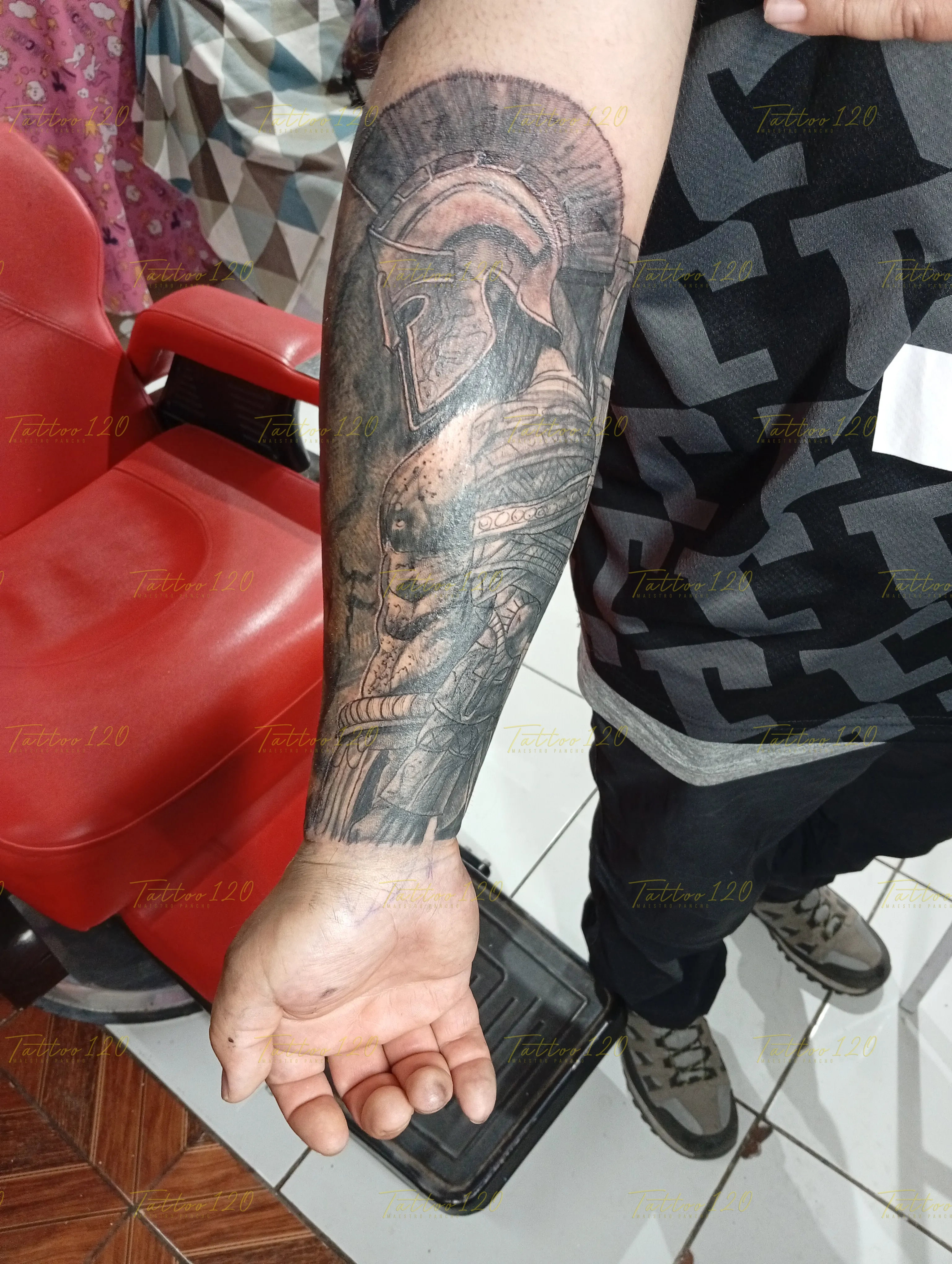 Tatuaje realista en el antebrazo de un guerrero espartano portando su casco clásico con cresta y armadura texturizada en tonos grises, en estilo de realistas, retratos y sombras, realizado en el estudio Tattoo120 en Colina, Región Metropolitana.