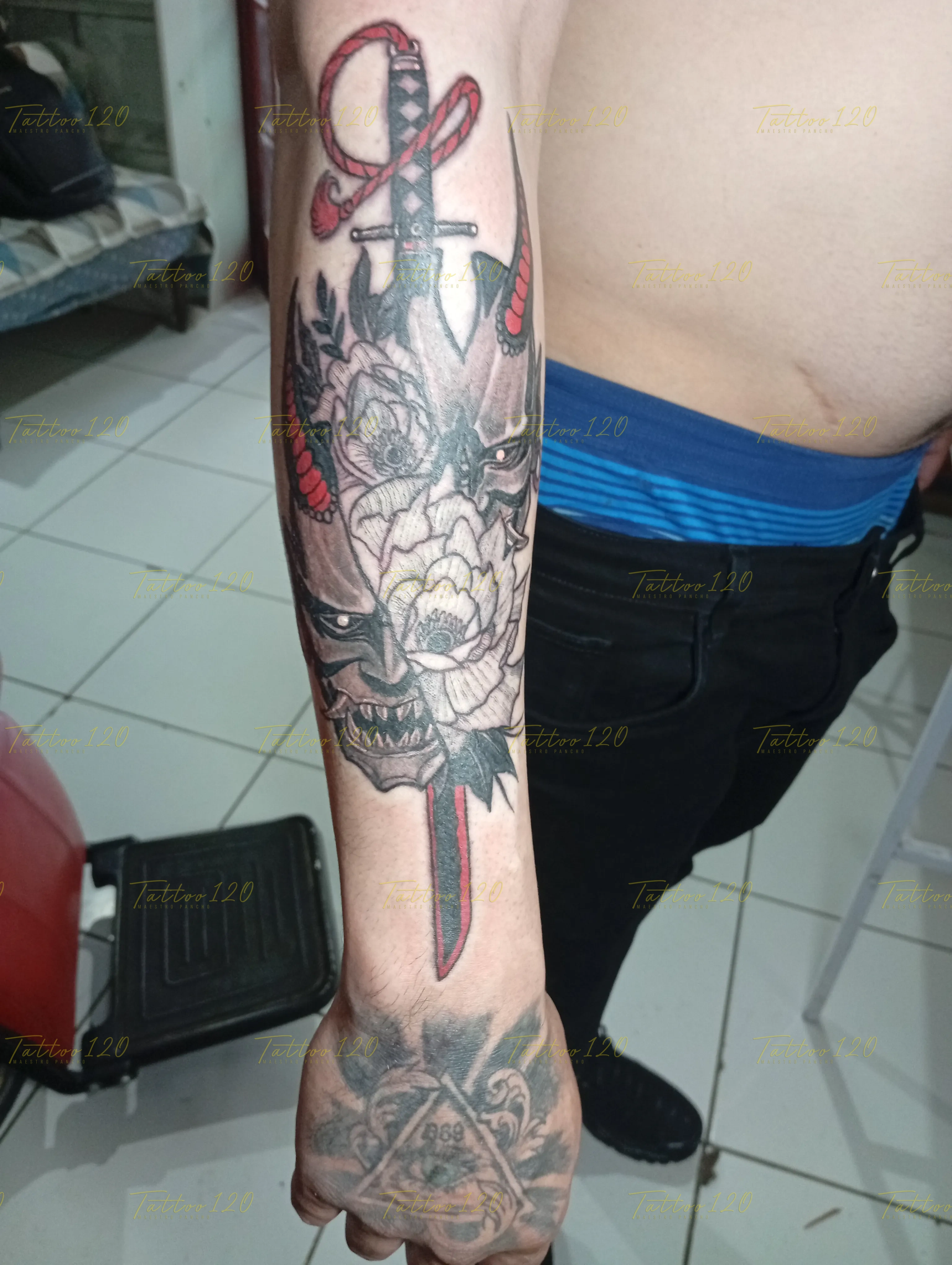 Tatuaje en el antebrazo de una espada japonesa que atraviesa una máscara Hannya dividida por flores de loto con sutiles toques rojos, en estilo de realistas, retratos y sombras, realizado en el estudio Tattoo120 en Colina, Región Metropolitana.