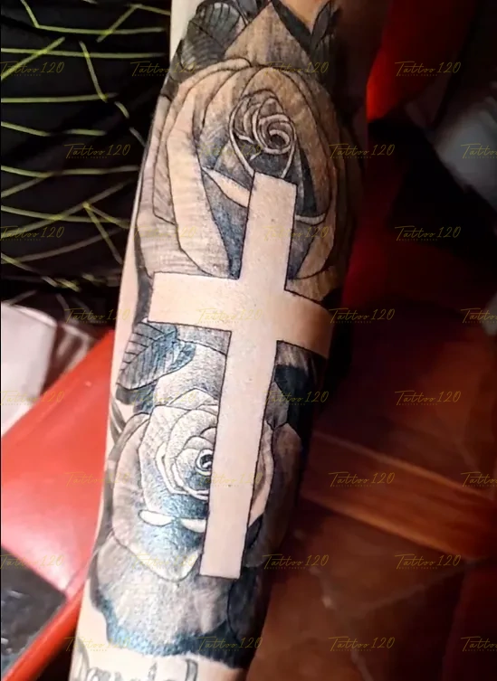Tatuaje en el brazo que destaca una cruz limpia en espacio negativo recortada sobre un fondo denso de rosas sombreadas, en estilo de realistas, retratos y sombras, realizado en el estudio Tattoo120 en Colina, Región Metropolitana.