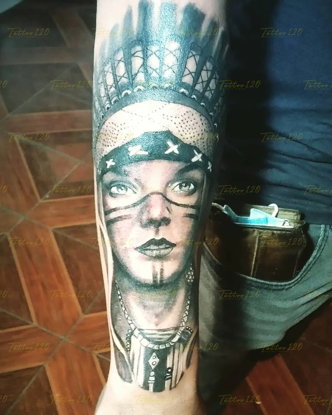 Tatuaje realista en el antebrazo del rostro de una mujer nativa con marcas en los ojos, pañuelo estampado y plumas en la cabeza, en estilo de realistas, retratos y sombras, realizado en el estudio Tattoo120 en Colina, Región Metropolitana.