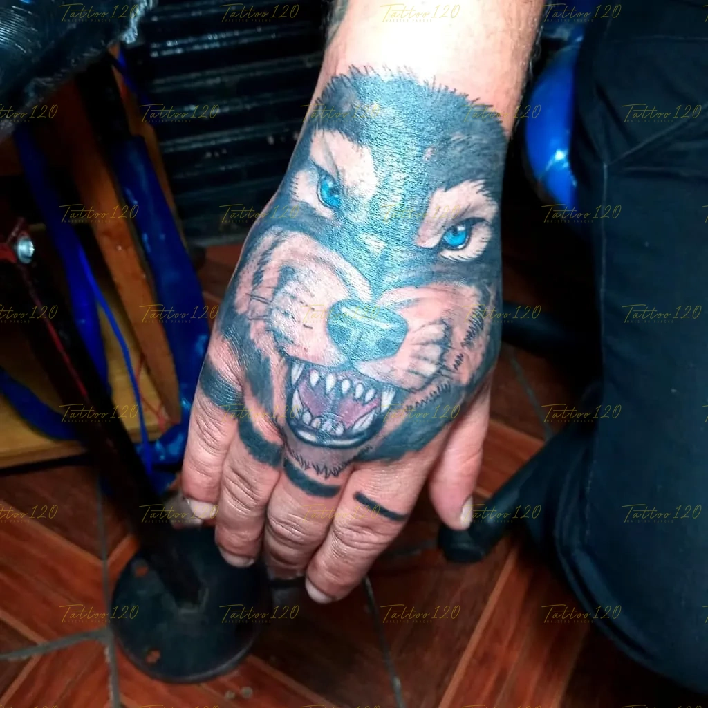 Tatuaje en el dorso de la mano que representa la cabeza de un lobo rugiendo con colmillos definidos y llamativos ojos de color azul, en estilo de realistas, retratos y sombras, realizado en el estudio Tattoo120 en Colina, Región Metropolitana.