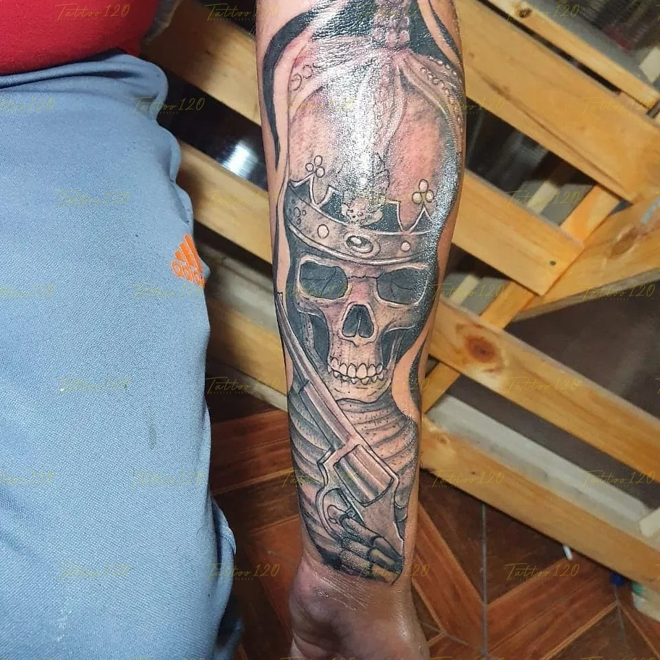 Tatuaje en el antebrazo que muestra una calavera con una corona ornamentada sosteniendo un revólver de estilo clásico con la mano ósea, en estilo de realistas, retratos y sombras, realizado en el estudio Tattoo120 en Colina, Región Metropolitana.