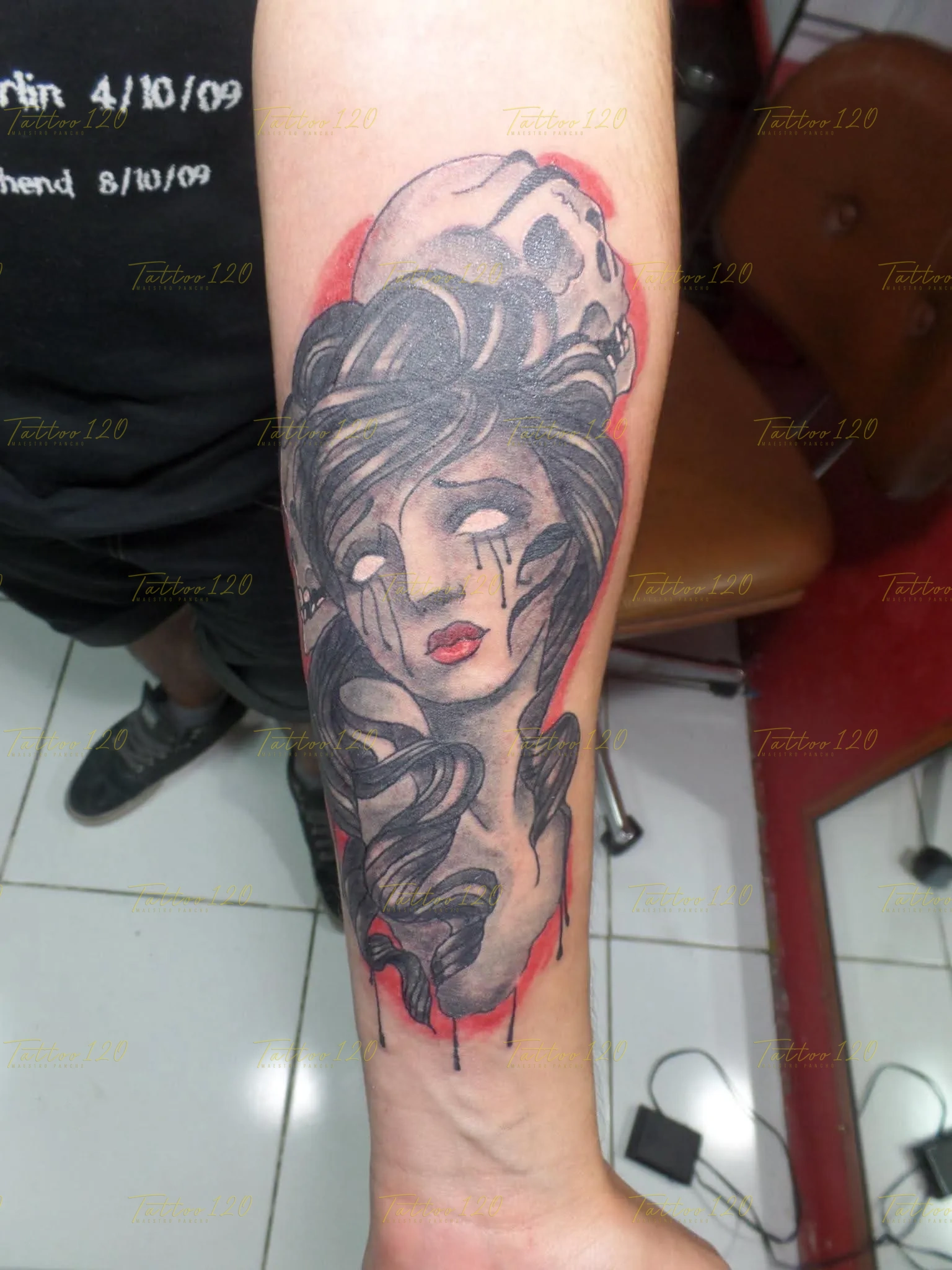 Tatuaje en el antebrazo que muestra el rostro de una mujer con lágrimas negras y ojos blancos bajo una calavera con fondo rojo, en estilo de tradicional y neo tradicional, realizado en el estudio Tattoo120 en Colina, Región Metropolitana.